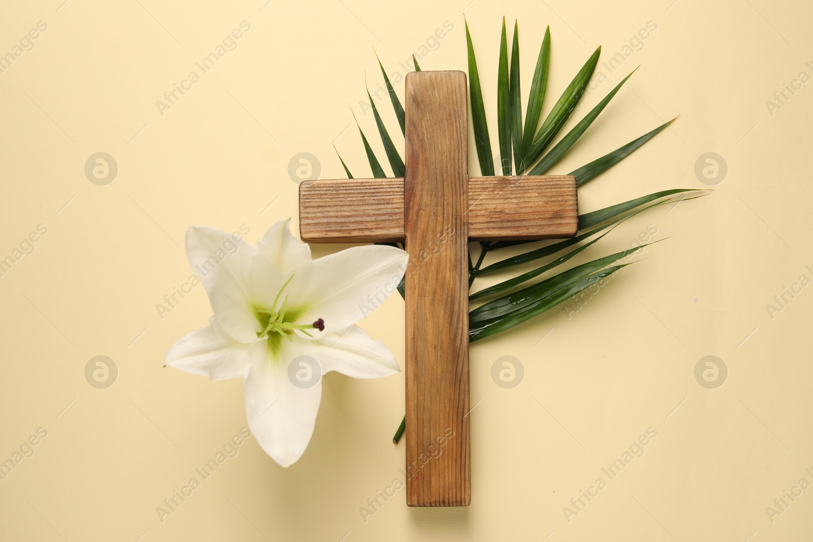 Photo of Wooden cross, lily flower and palm leaf on pale yellow background, top view. Easter attributes