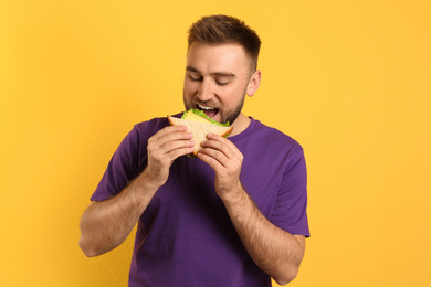 Young man eating tasty sandwich on yellow background