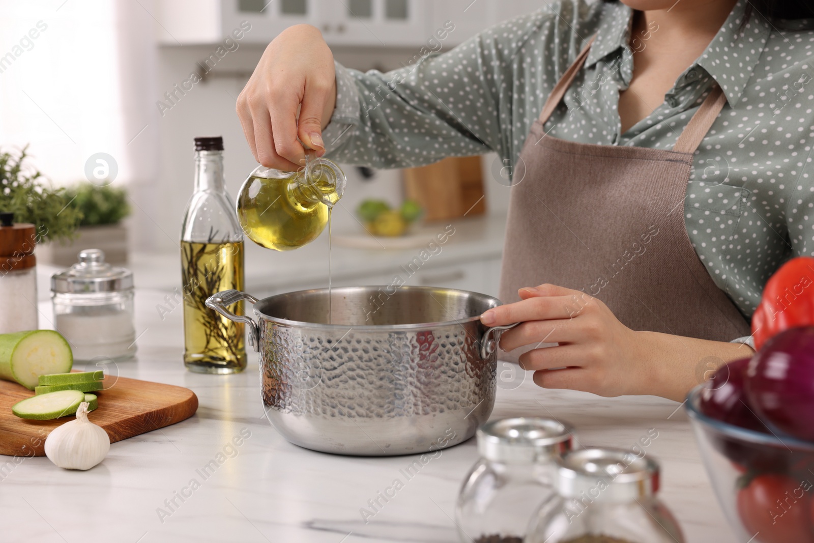 Photo of Cooking process. Woman pouring oil from bottle into pot at white countertop in kitchen, closeup