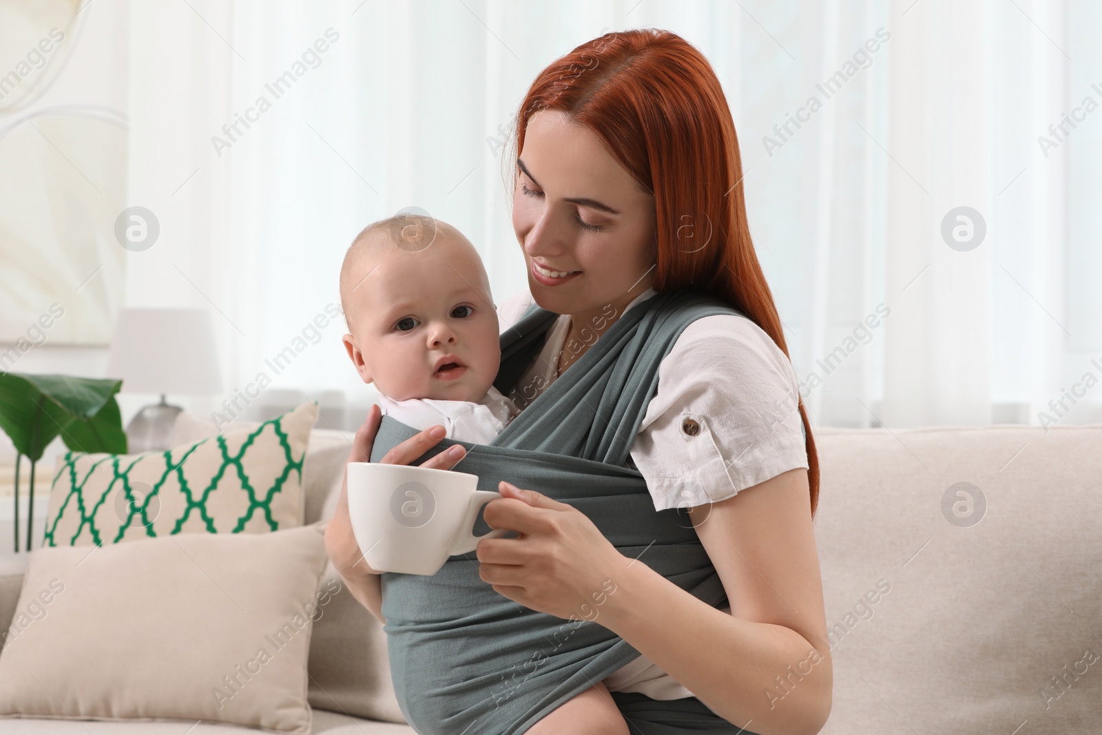 Photo of Mother with cup of drink holding her child in sling (baby carrier) at home