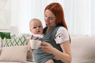 Photo of Mother with cup of drink holding her child in sling (baby carrier) at home