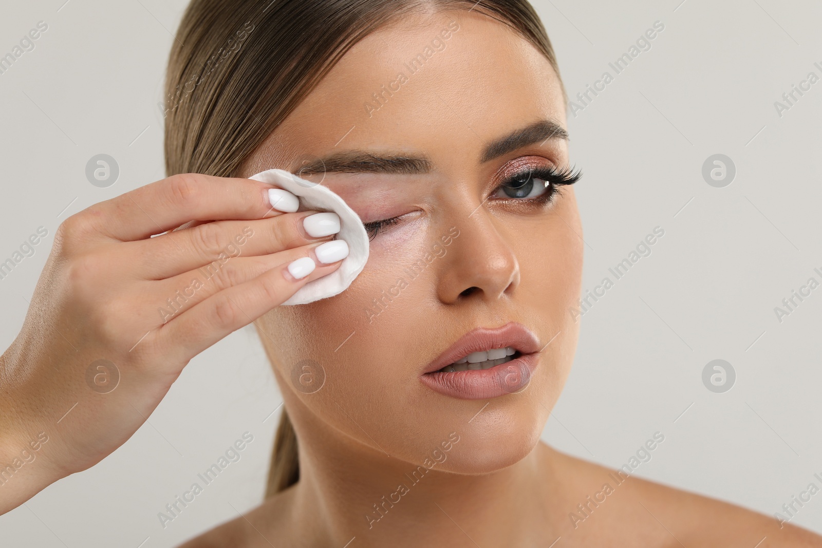 Photo of Beautiful woman removing makeup with cotton pad on light grey background, closeup