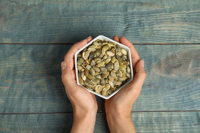 Photo of Young woman with bowl of raw pumpkin seeds at blue wooden table, top view