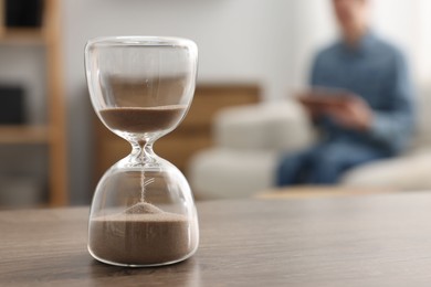 Photo of Hourglass with flowing sand on desk. Man reading book in room, selective focus