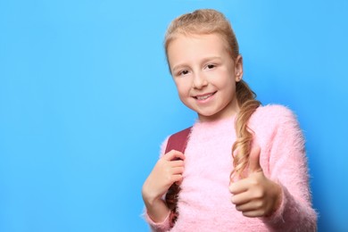 Photo of Happy little girl with backpack showing thumbs up on light blue background. Space for text