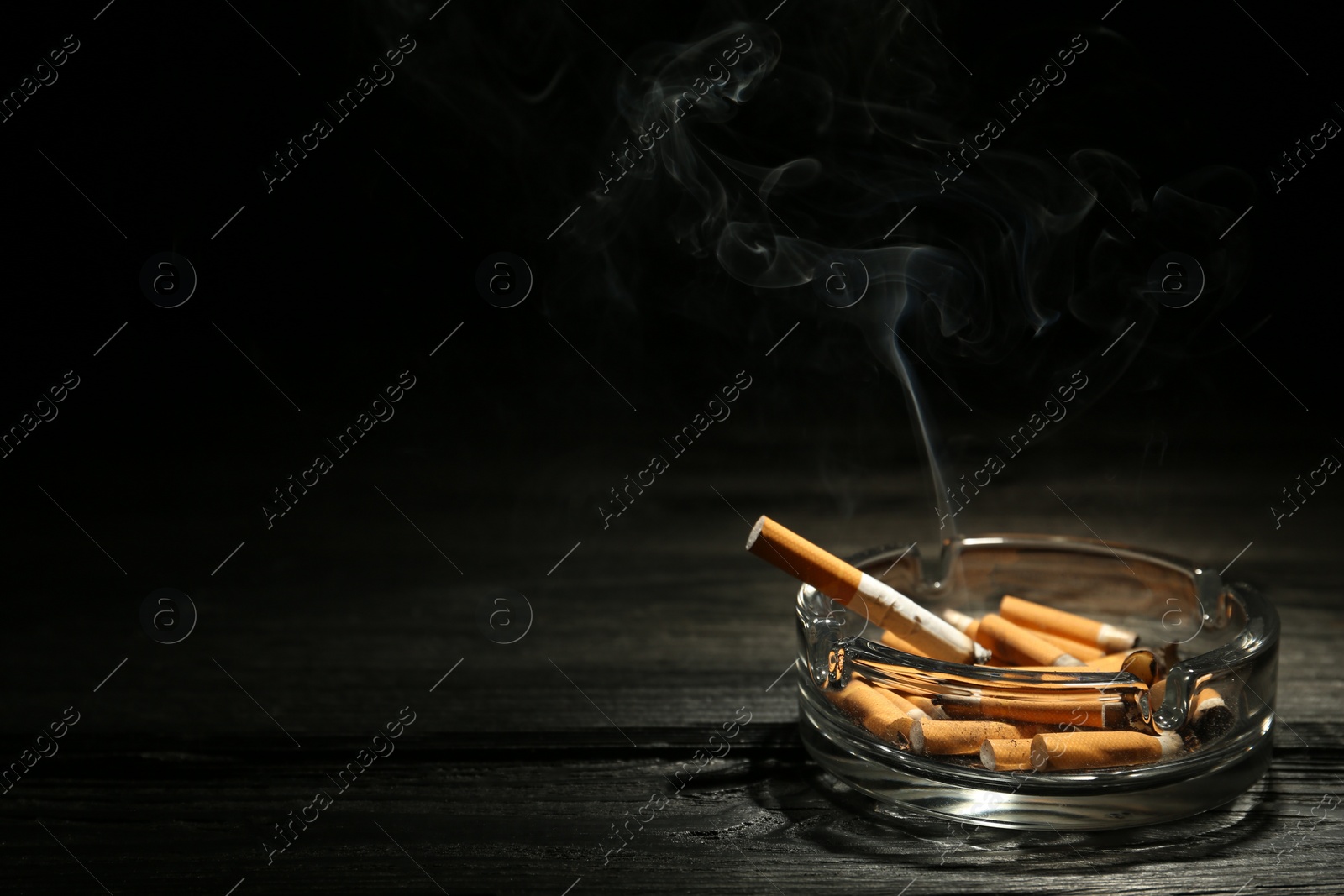 Photo of Smoldering cigarette in glass ashtray on dark wooden table against black background. Space for text