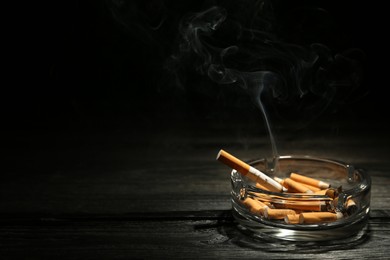Photo of Smoldering cigarette in glass ashtray on dark wooden table against black background. Space for text