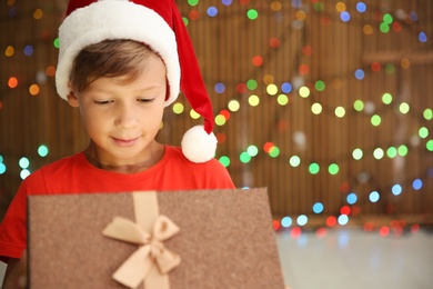Cute little child in Santa hat opening Christmas gift box on blurred lights background