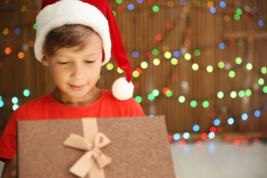 Photo of Cute little child in Santa hat opening Christmas gift box on blurred lights background