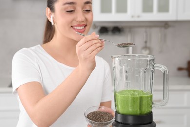 Photo of Beautiful young woman adding chia seeds into delicious smoothie in kitchen, focus on blender