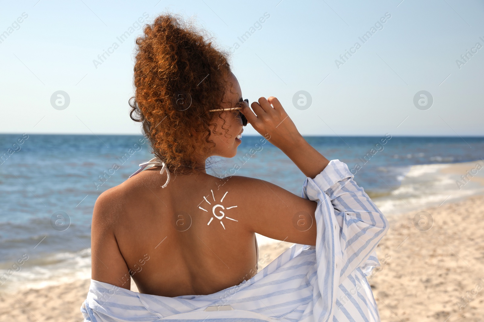Photo of Beautiful African American woman with sun protection cream on shoulder at beach, back view
