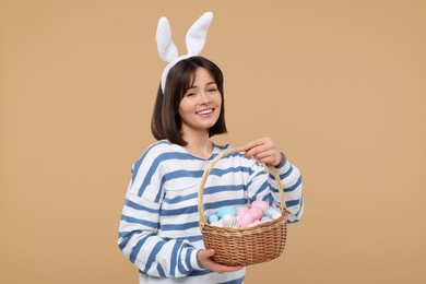 Photo of Easter celebration. Happy woman with bunny ears and wicker basket full of painted eggs on beige background