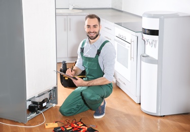 Photo of Male technician with clipboard and tools near broken refrigerator in kitchen