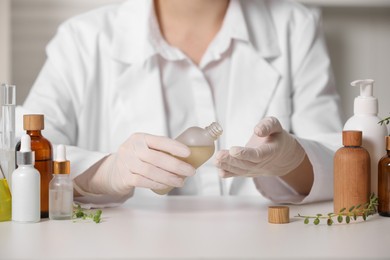 Photo of Dermatologist with bottle testing cosmetic product at white table indoors, closeup