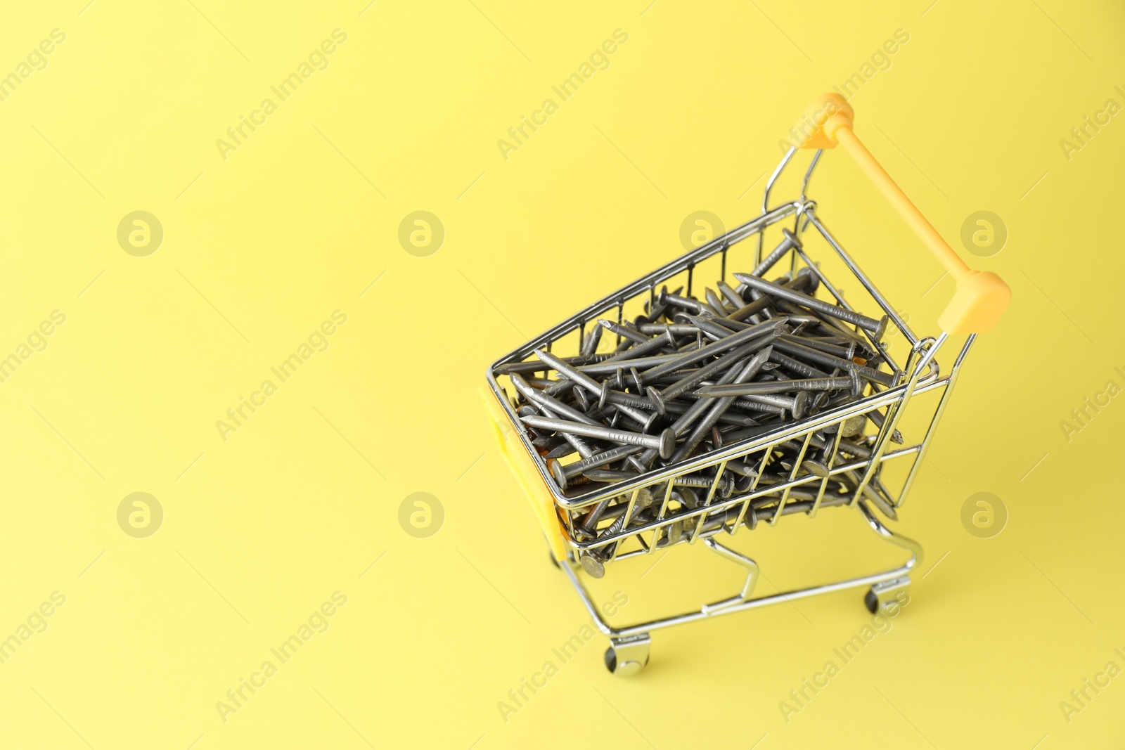 Photo of Metal nails in shopping cart on yellow background, space for text