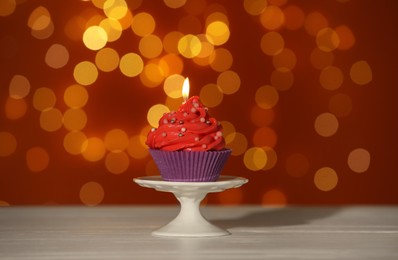 Photo of Stand with birthday cupcake on white table against blurred lights