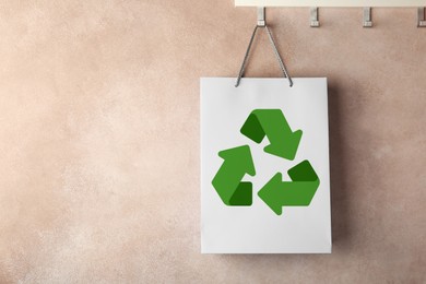 Paper shopping bag with recycling symbol hanging on beige wall. Space for text