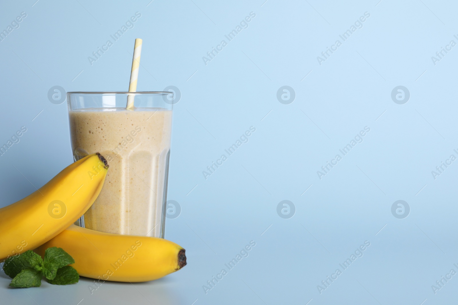 Photo of Glass of tasty smoothie with straw, bananas and mint leaves on light blue background. Space for text