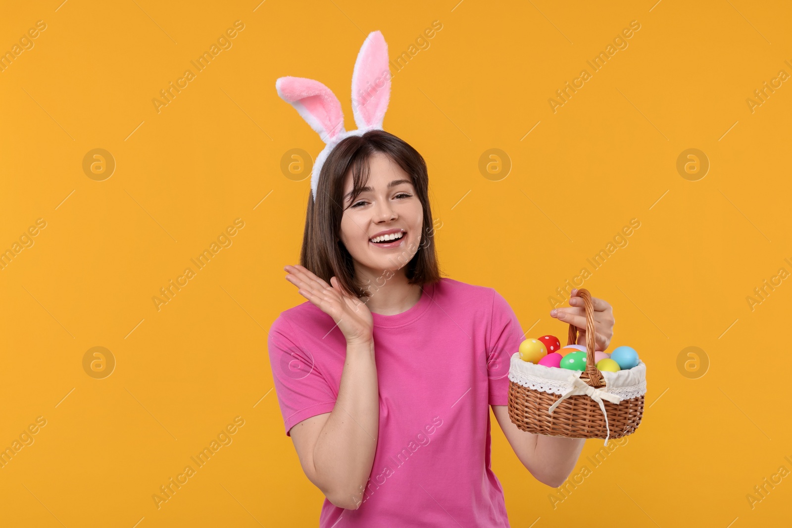 Photo of Easter celebration. Happy woman with bunny ears and wicker basket full of painted eggs on orange background