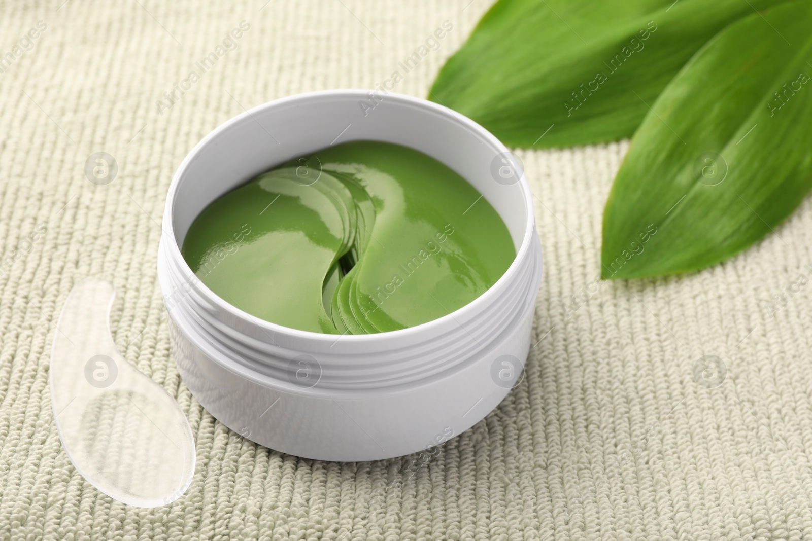 Photo of Jar of under eye patches with spoon and green leaves on light cloth, closeup. Cosmetic product