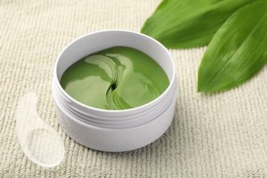Photo of Jar of under eye patches with spoon and green leaves on light cloth, closeup. Cosmetic product