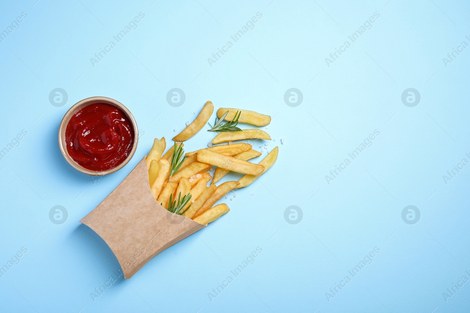 Photo of Paper cup with French fries, rosemary and ketchup on light blue table, flat lay. Space for text