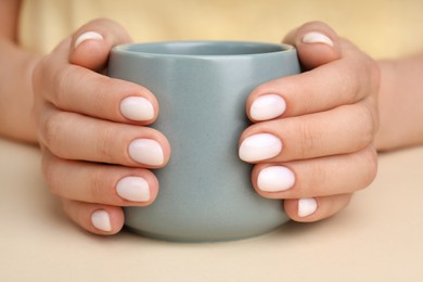 Photo of Woman with white nail polish holding cup at beige table, closeup