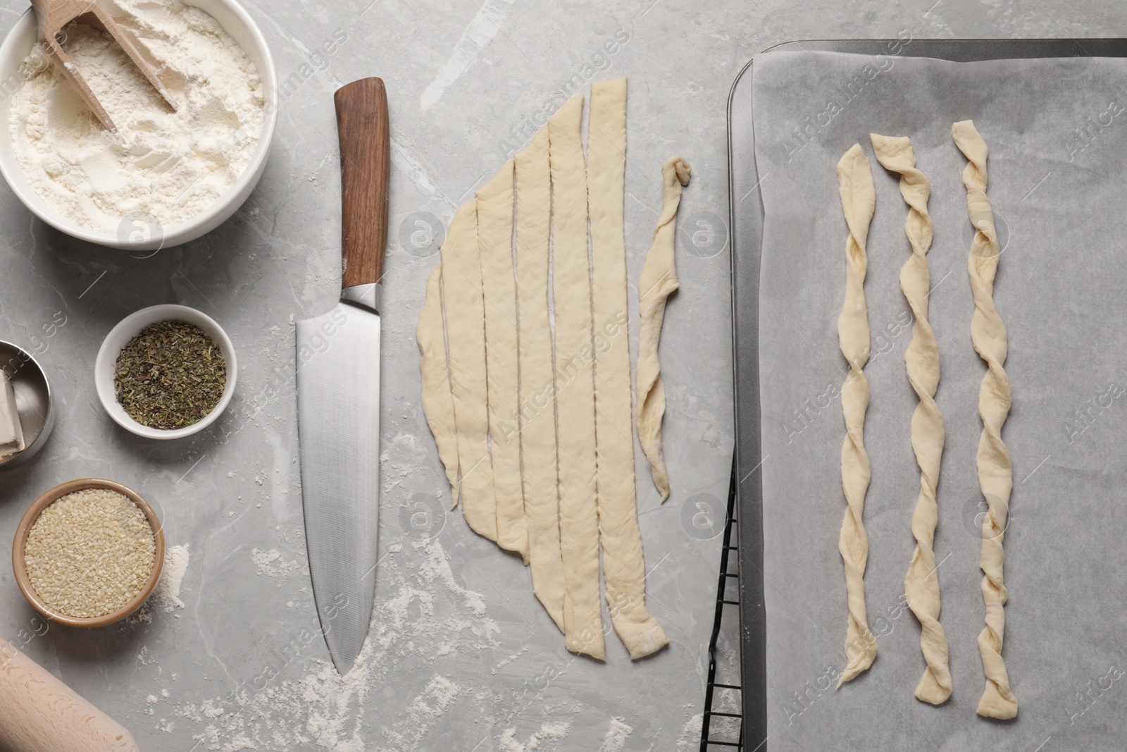 Photo of Baking sheet with homemade breadsticks, knife and ingredients on grey marble table, flat lay. Cooking traditional grissini