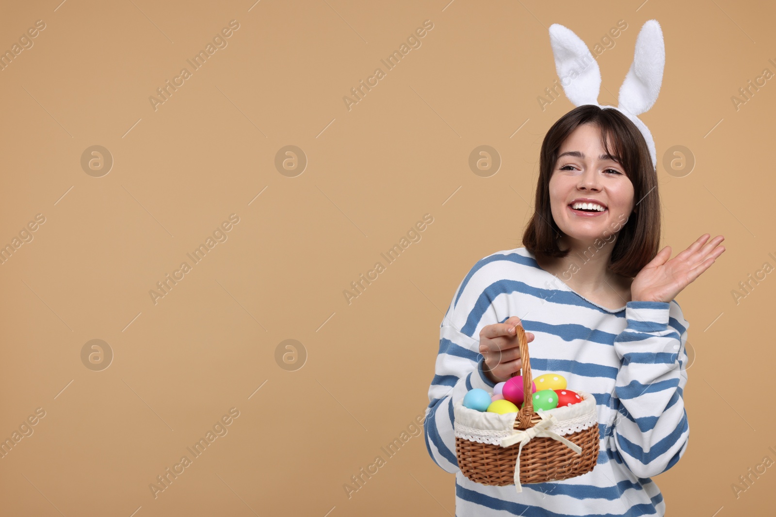 Photo of Easter celebration. Happy woman with bunny ears and wicker basket full of painted eggs on beige background, space for text