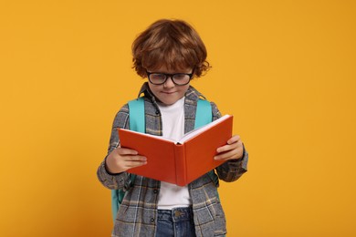 Happy schoolboy with backpack reading book on orange background