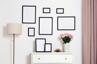 Empty frames hanging on white wall and chest of drawers with flowers indoors