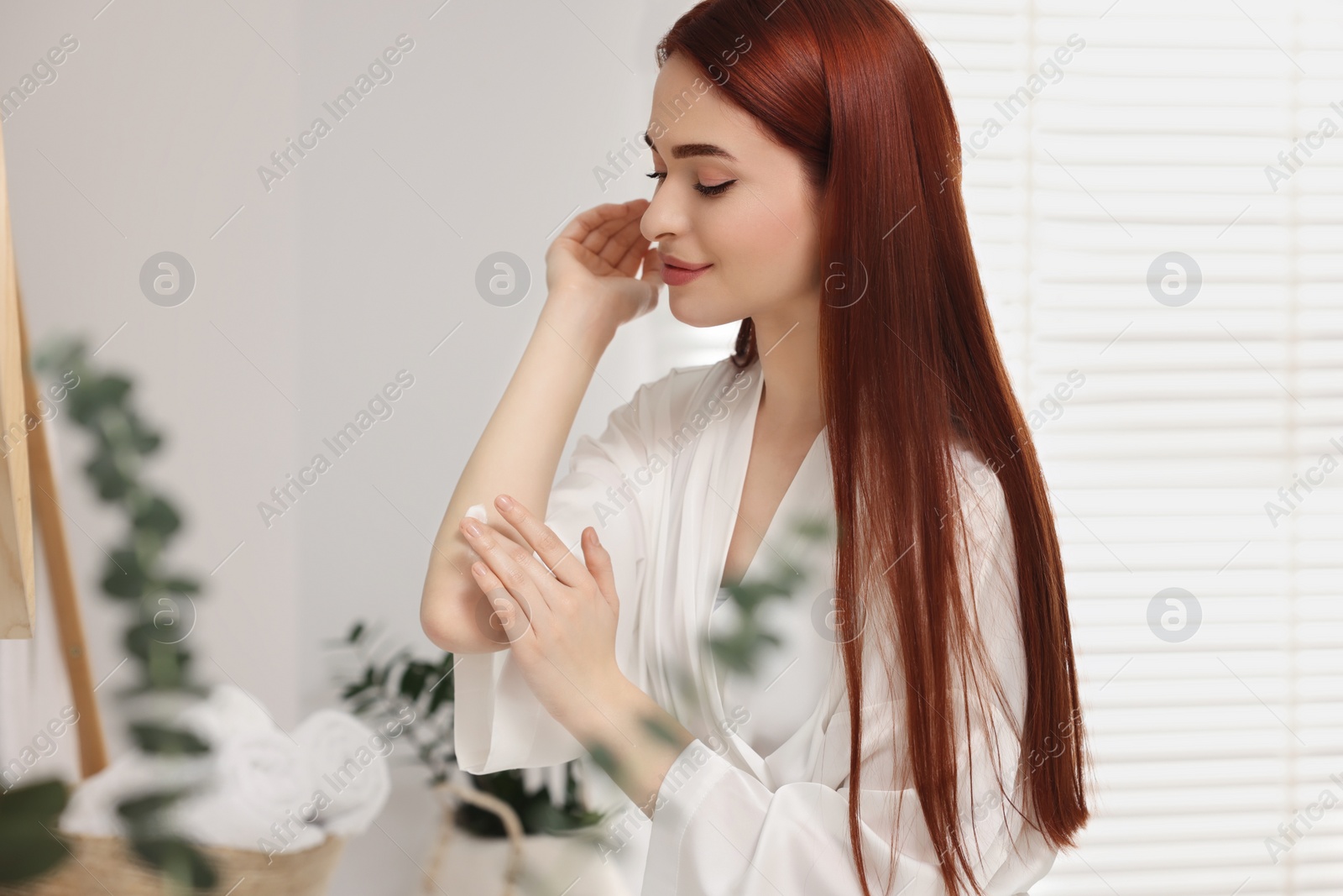 Photo of Beautiful young woman applying body cream onto elbow in bathroom, space for text