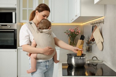 Photo of Mother holding her child in sling (baby carrier) while cooking pasta in kitchen