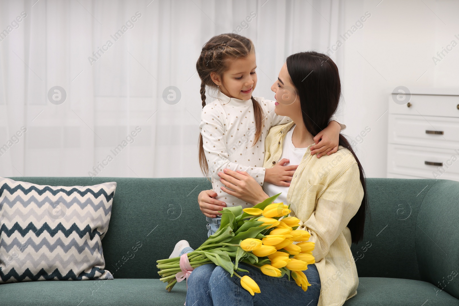 Photo of Happy woman with her daughter and bouquet of yellow tulips on sofa at home, space for text. Mother's day celebration