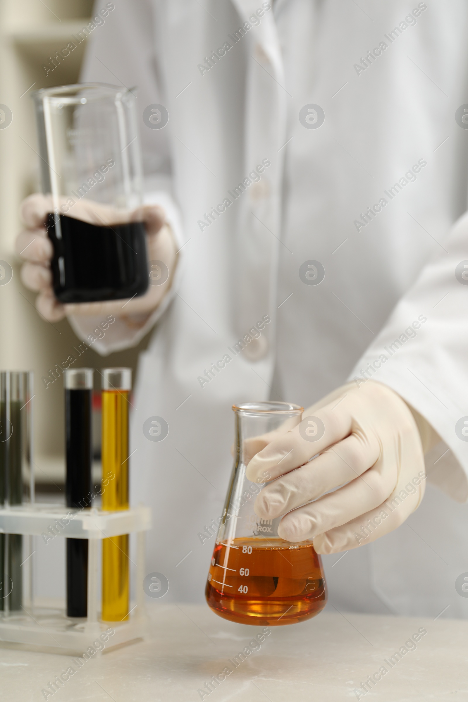 Photo of Laboratory worker holding beaker and flask with different types of crude oil at light marble table, closeup