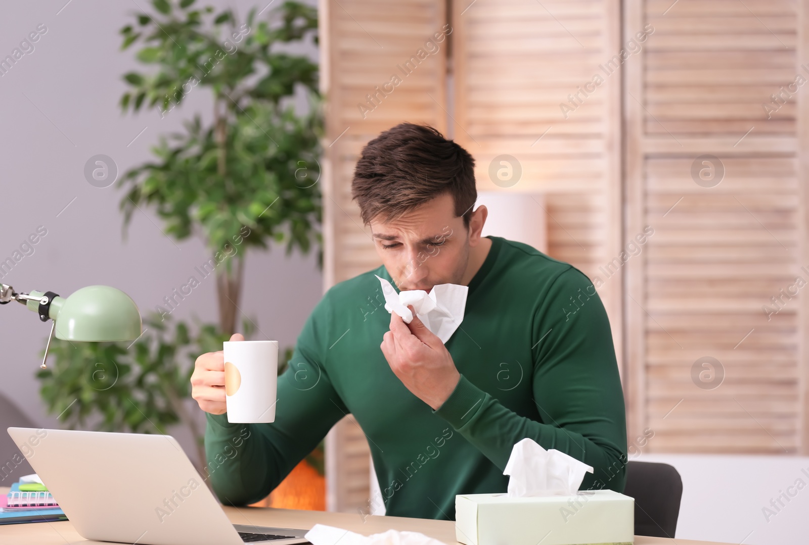 Photo of Sad exhausted man with tissue and cup of hot drink suffering from cold while working with laptop at table