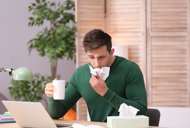 Photo of Sad exhausted man with tissue and cup of hot drink suffering from cold while working with laptop at table