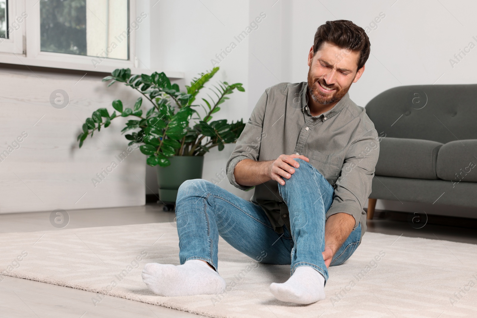 Photo of Man suffering from leg pain on carpet near sofa at home