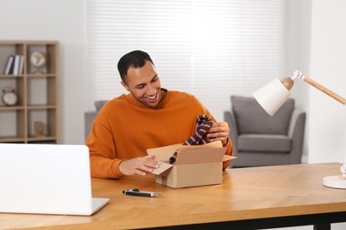 Photo of Happy young man opening parcel at table indoors. Internet shopping