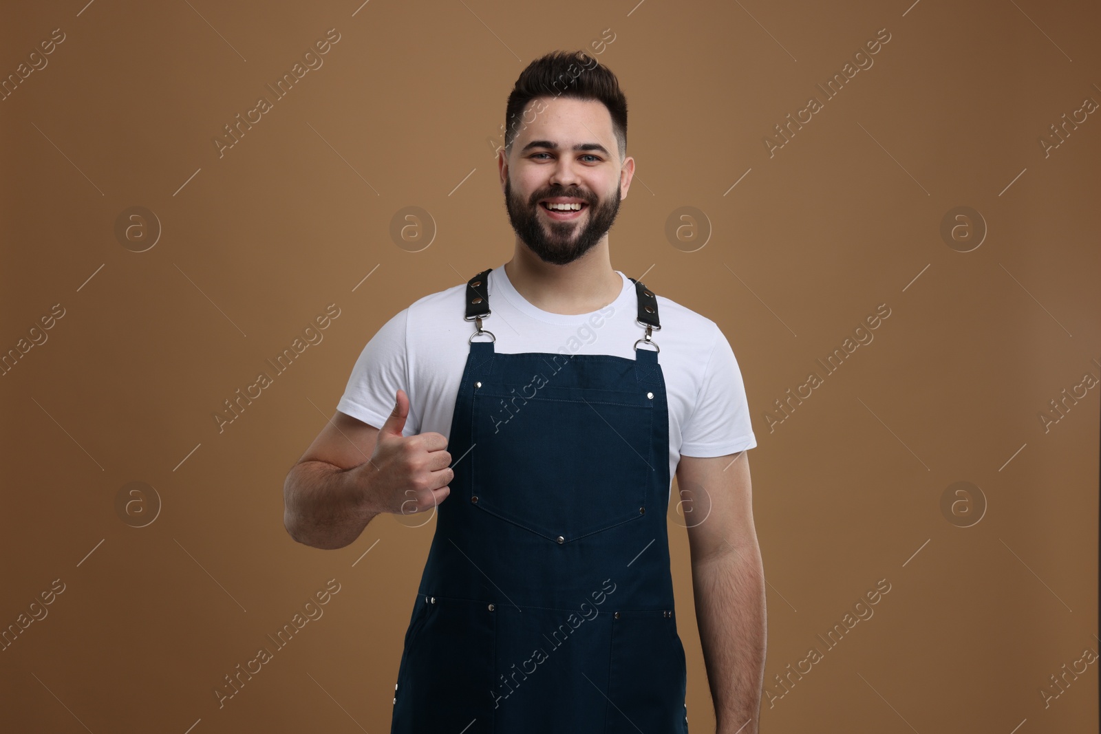 Photo of Smiling man in kitchen apron showing thumb up on brown background. Mockup for design
