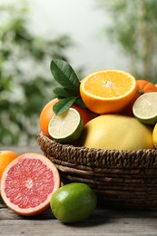 Different fresh citrus fruits and leaves in wicker basket on wooden table against blurred background, closeup