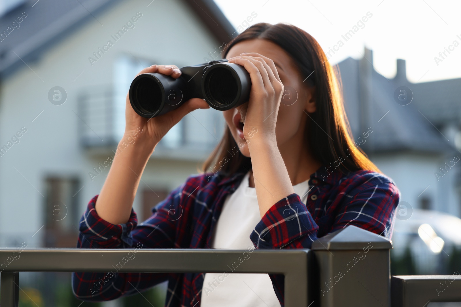 Photo of Concept of private life. Curious young woman with binoculars spying on neighbours over fence outdoors