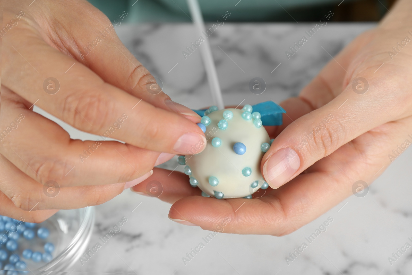 Photo of Young woman decorating tasty cake pop with blue sprinkles at white marble table, closeup