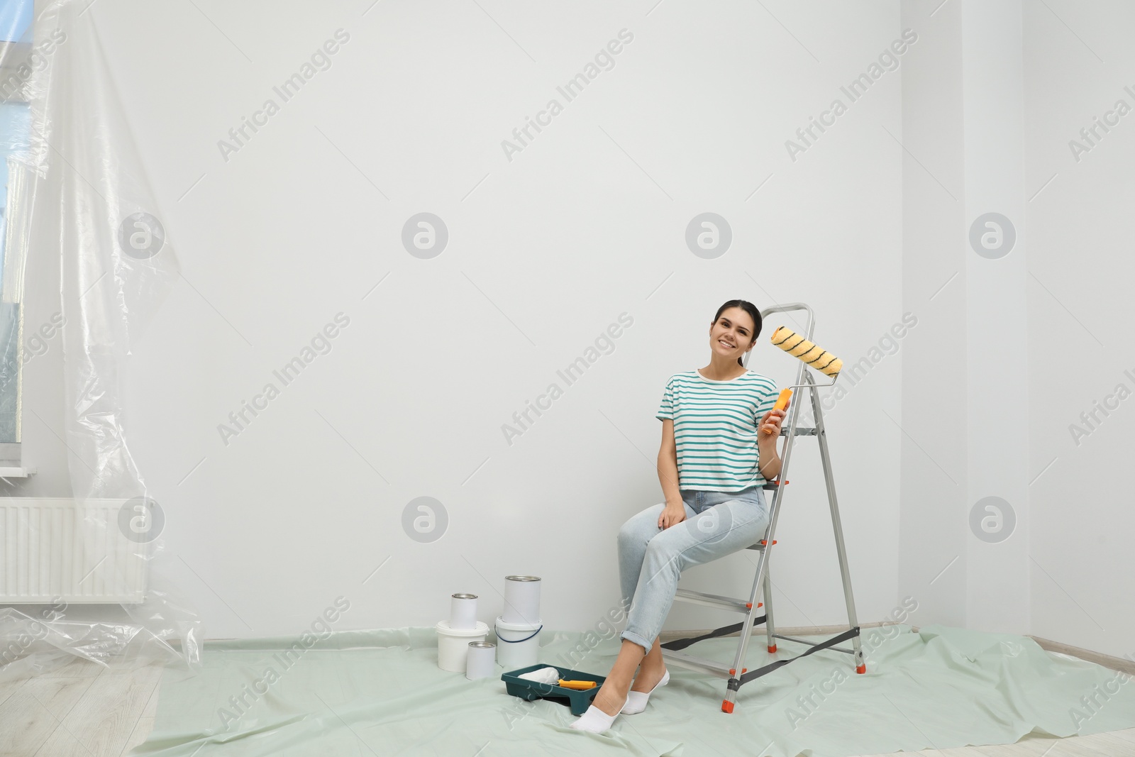 Photo of Woman with paint roller brush sitting on metallic folding ladder indoors, space for text