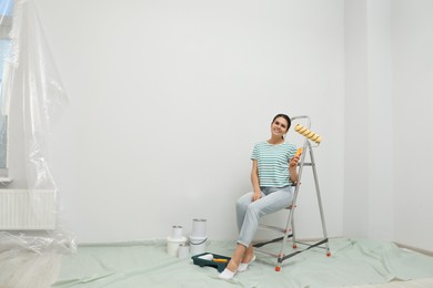 Photo of Woman with paint roller brush sitting on metallic folding ladder indoors, space for text