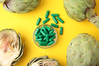 Photo of Bowl with pills and fresh artichokes on yellow background, flat lay
