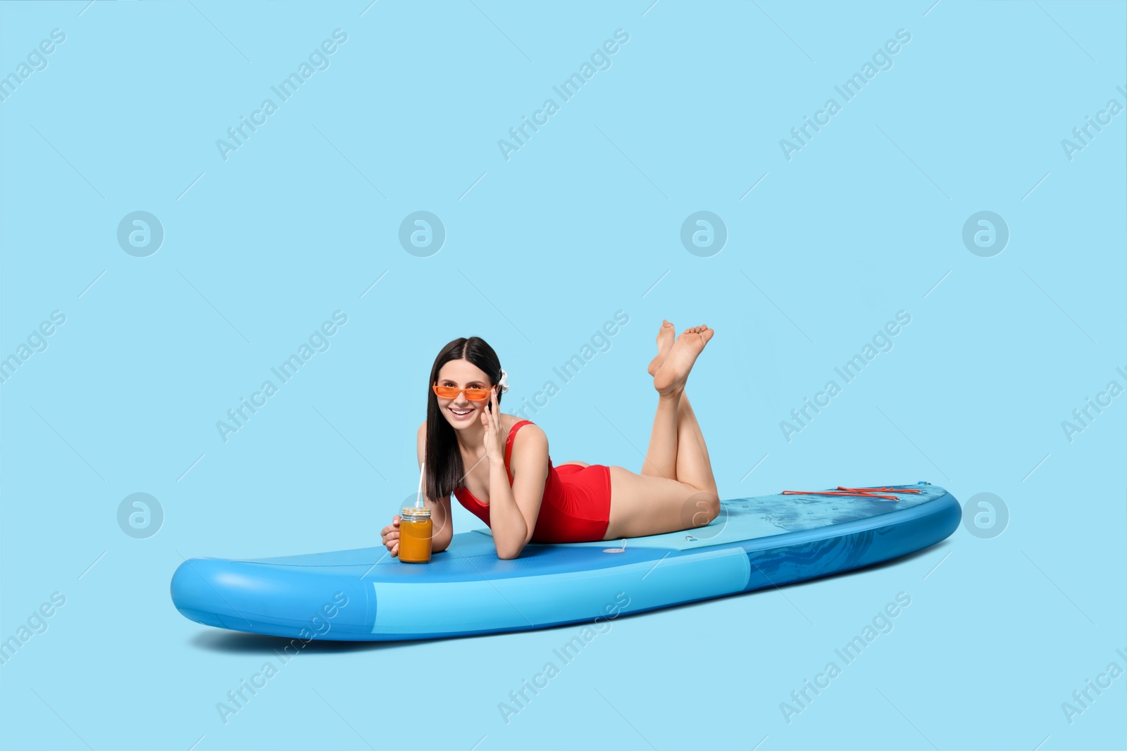 Photo of Happy woman with refreshing drink resting on SUP board against light blue background