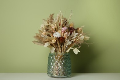 Photo of Beautiful dried flower bouquet in glass vase on white table near olive wall