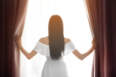 Woman opening window curtains at home, back view