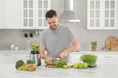 Happy man cutting celery for delicious smoothie at white marble table in kitchen